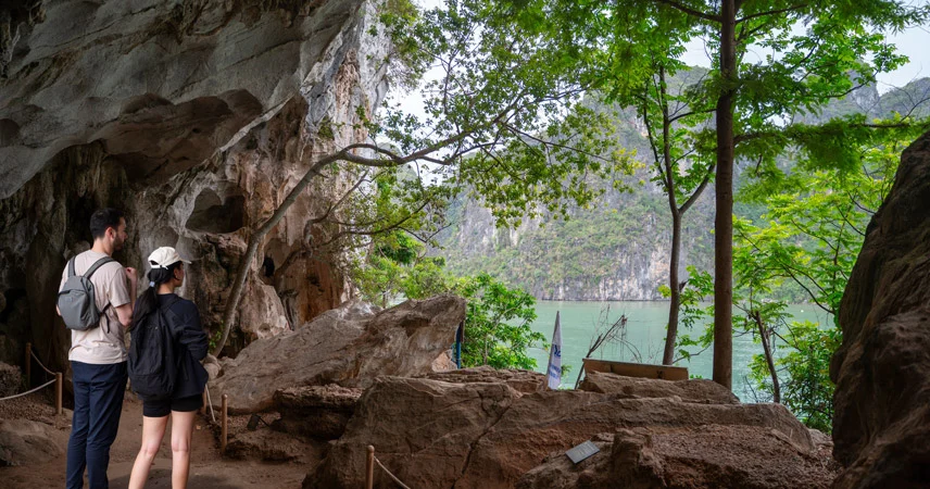 Two visitors look toward green bay waters from the rocky entrance of Thien Canh Son Cave.