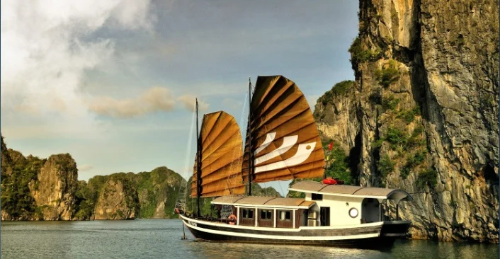 A traditional junk with brown sails moves beside a limestone cliff.