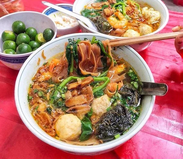 Bowls of banh da cua contain brown rice noodles, greens and assorted toppings on a red table.