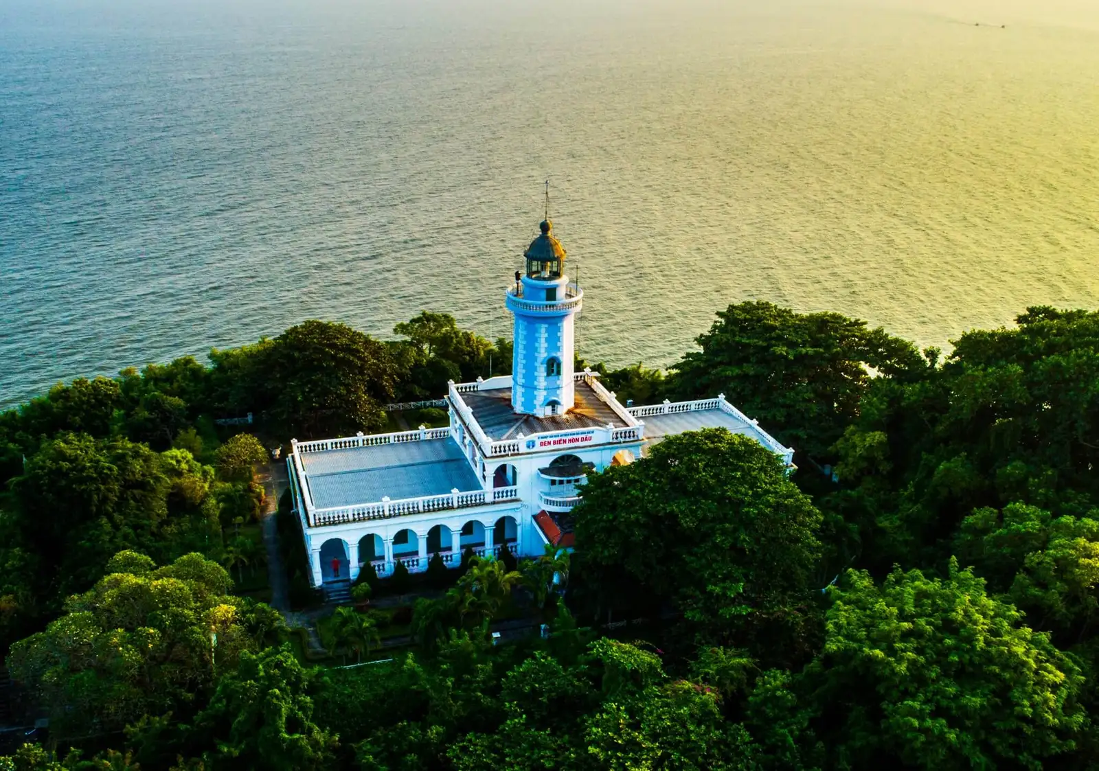 Hon Dau Lighthouse rises above a wooded island beside the sea.