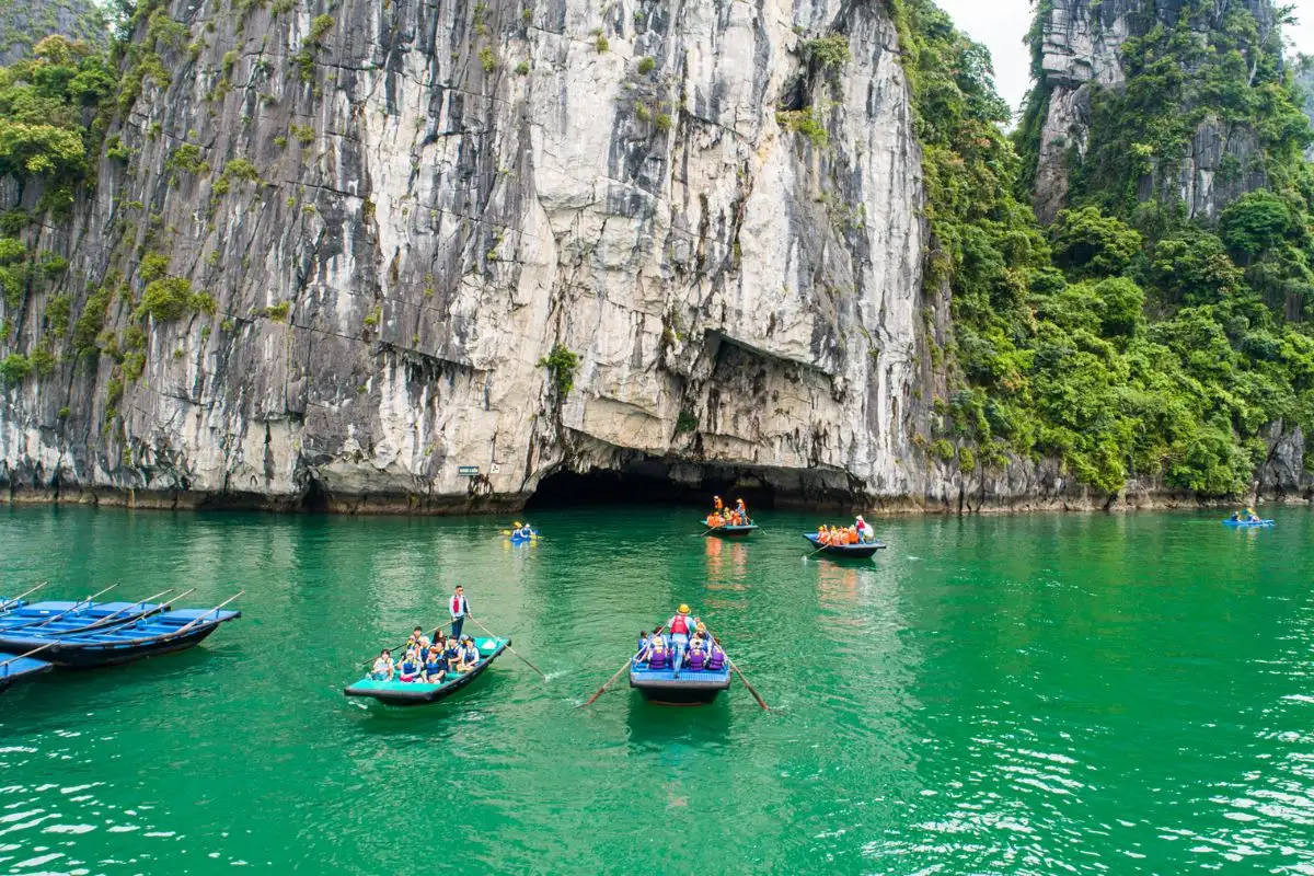 Small boats and kayaks gather on green water below a limestone cliff.