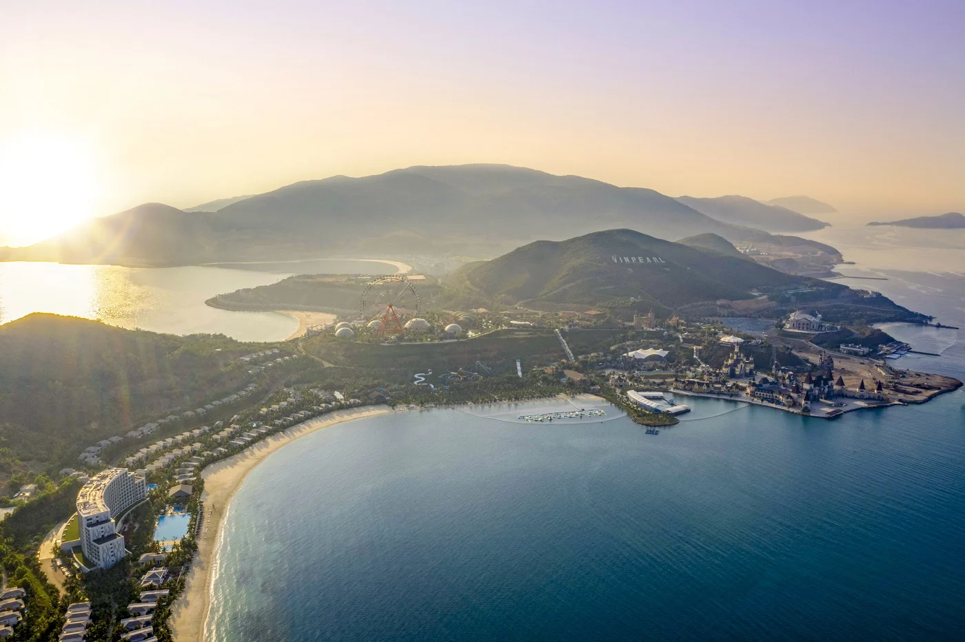 An aerial view shows hills, resort buildings and a curving bay on Hon Tre Island at sunrise.